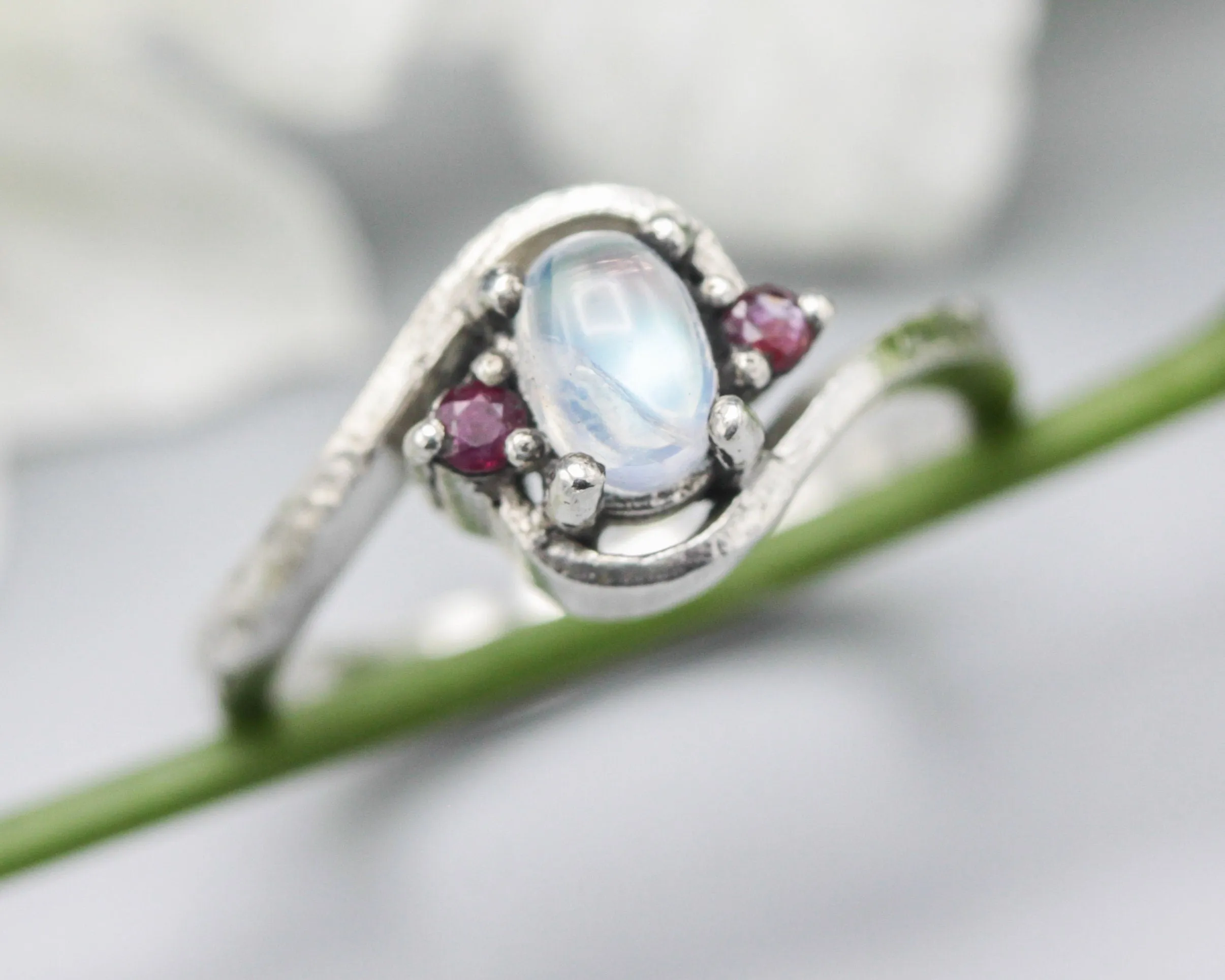 Oval cabochon moonstone ring with tiny ruby side set gems in prongs setting with sterling silver oxidized texture band