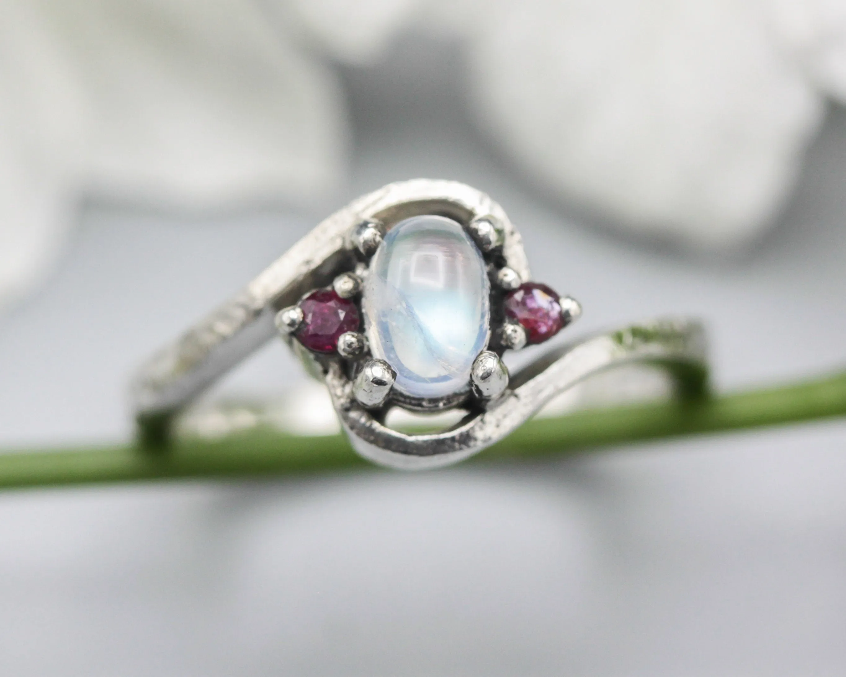 Oval cabochon moonstone ring with tiny ruby side set gems in prongs setting with sterling silver oxidized texture band